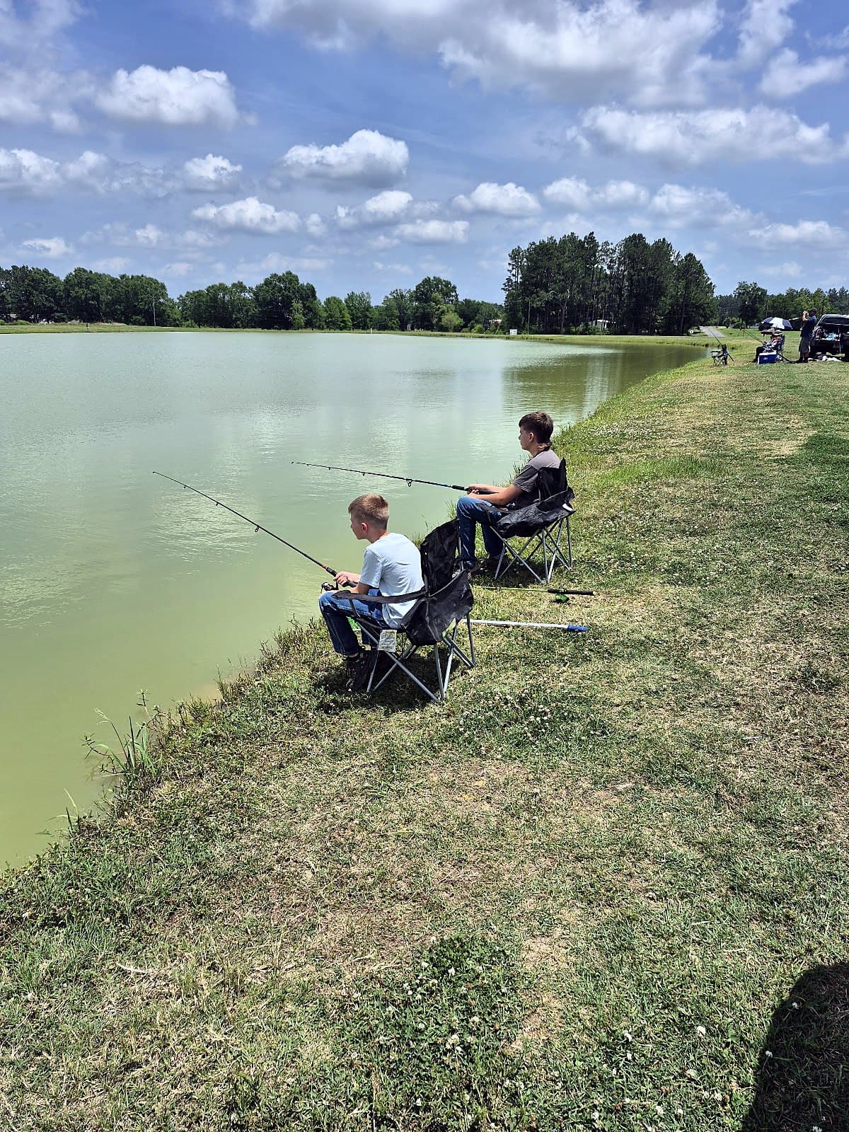 Anglers fishing along the grassy bank of a stocked pond under a blue sky at Sunrise Catfish Farm